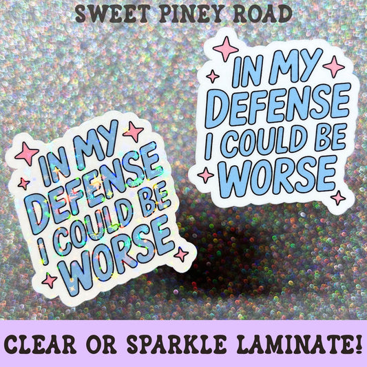 Funny pastel blue sticker featuring the phrase In My Defense I Could Be Worse with pink star accents and a playful sarcastic design.