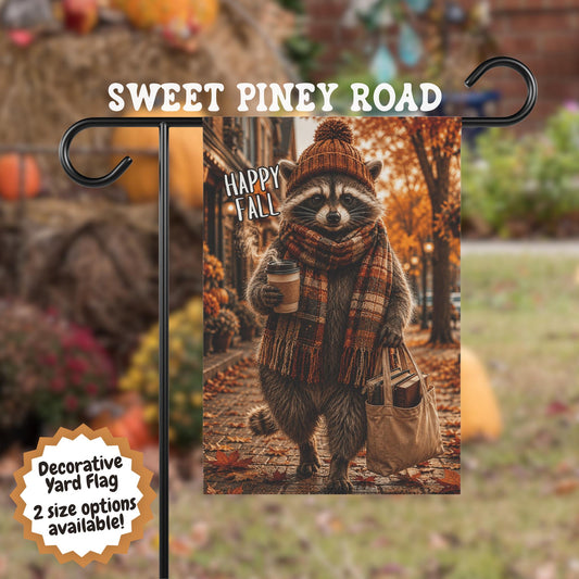 Happy Fall yard flag featuring a raccoon wearing a knit beanie and plaid scarf, holding a coffee cup and tote bag filled with books while walking through a colorful autumn street lined with fall leaves.