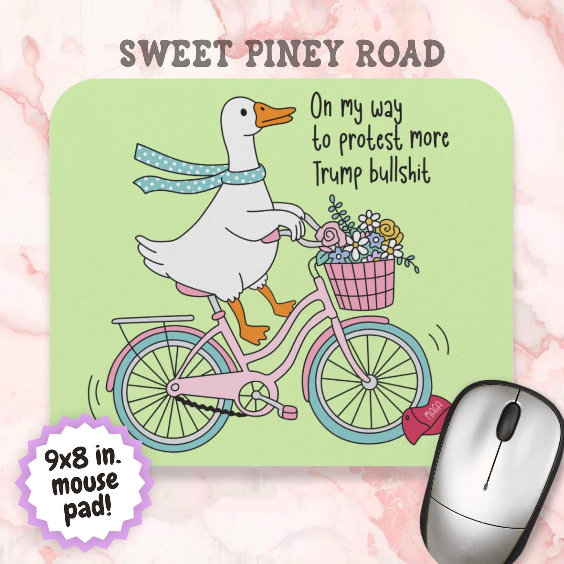 Funny anti-Trump mouse pad featuring a white goose riding a pink bicycle with a basket of flowers and a blue scarf. The artwork includes the phrase "On my way to protest more Trump bullshit" and a red MAGA hat on the ground, combining political satire with pastel cottagecore aesthetics.