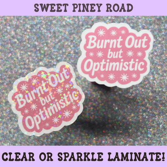 Pink die-cut sticker reading "Burnt Out But Optimistic" with retro typography and star accents on a white background.