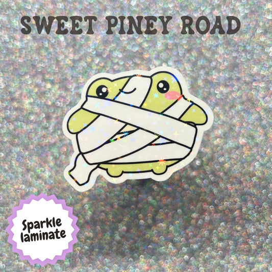 Cute kawaii toad wrapped in mummy bandages with a smiling face, featuring a playful Halloween design and displayed with a sparkly laminate finish.