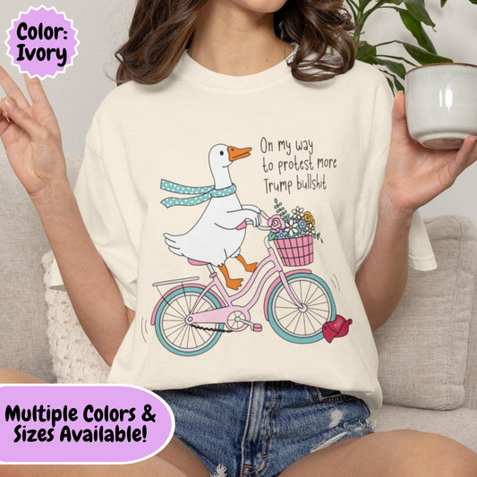 Cartoon goose riding a pink bicycle with a flower basket while running over a red MAGA hat, colorful political humor graphic featuring whimsical artwork and anti-MAGA satire.