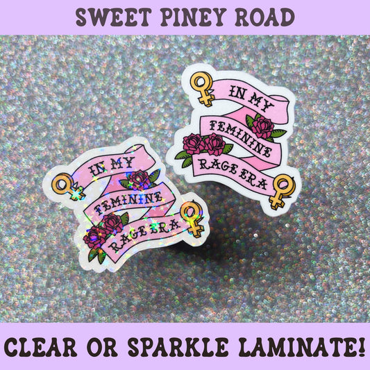 Feminist sticker featuring pink banners reading "In My Feminine Rage Era" with roses and female symbols on holographic sparkle laminate.